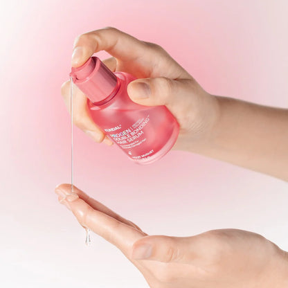 Hand holding a pink serum bottle with a droplet of serum on a pink background